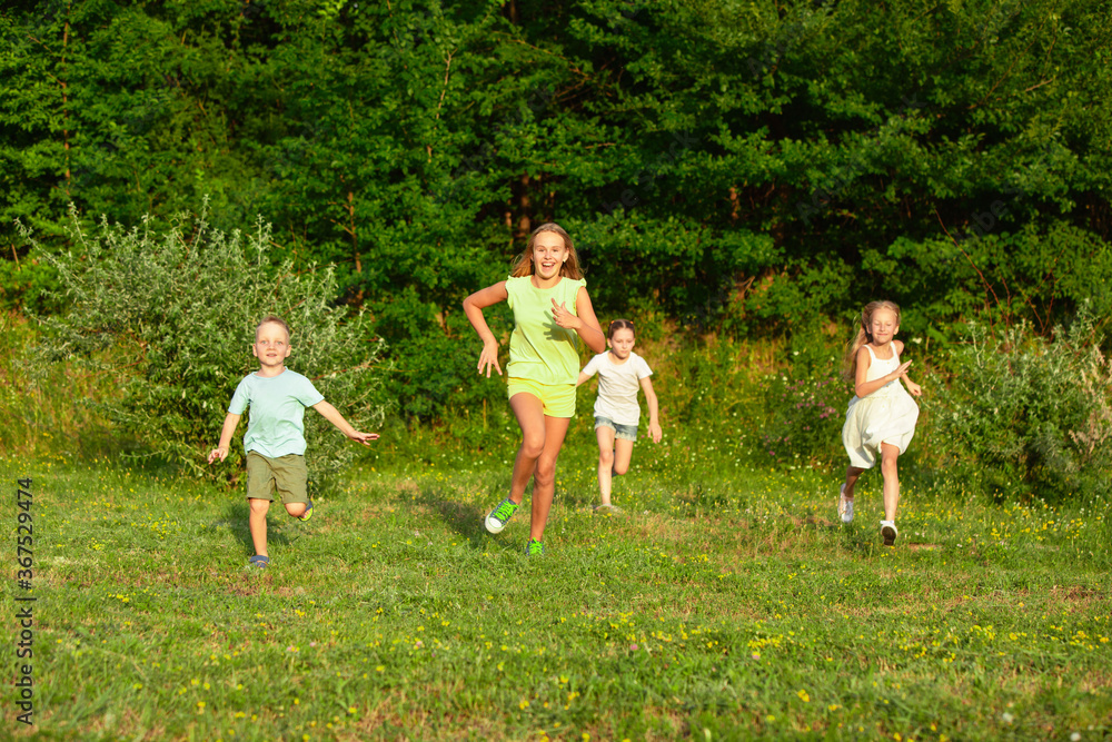 Fototapeta premium Kids, children running on meadow in summer's sunlight. Look happy, cheerful with sincere bright emotions. Cute caucasian boys and girls. Concept of childhood, happiness, movement, family and summer.