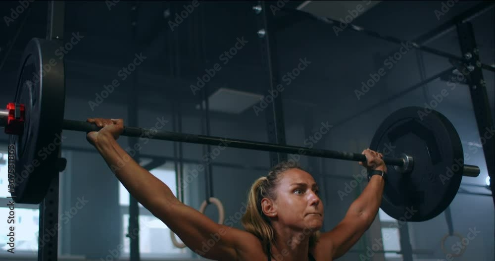 Slow motion: Training squats with barbells over head. Muscular female lifting barbell over head training shoulders at gym. Confident fitness woman practicing force workout with heavy equipment. Slow motion: Training squats with barbells over head. Muscular female lifting barbell over head training shoulders at gym. Confident fitness woman practicing force workout with heavy equipment.
