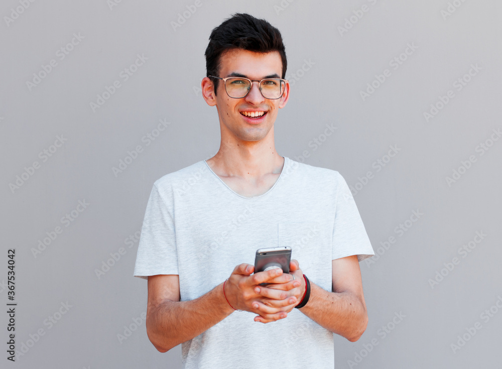 Young man leaning against a grey wall using mobile phone. Portrait of a happy laughing guy texting a phone message and looking at camera. Man reading a message on cell phone with copyspace. Young man leaning against a grey wall using mobile phone. Portrait of a happy laughing guy texting a phone message and looking at camera. Man reading a message on cell phone with copyspace.