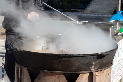 Cooking real Uzbek pilaf. Stewing lamb ribs in a huge cauldron with lots of steam outside during a traditional folk festival