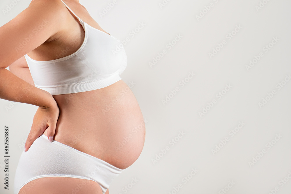 Close up of pregnant woman wearing supportive seamless maternity bra and maxi bottoms, arms on her belly. Female hands wrapped around big bare tummy. Child expectancy concept. Background, copy space Close up of pregnant woman wearing supportive seamless maternity bra and maxi bottoms, arms on her belly. Female hands wrapped around big bare tummy. Child expectancy concept. Background, copy space
