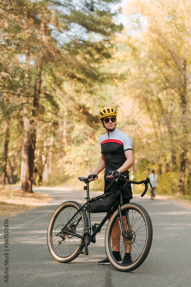 Cyclist in a sports outfit poses for a camera on the road in an autumn forest, stands with a bicycle rider and looks away with serious face. Concentrated young man with a bicycle relaxing on park