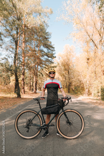 Portrait of young man with bicycle standing on road in autumn forest and posing at camera. Man is walking on a bicycle ride outside the city, standing against a beautiful autumn landscape.