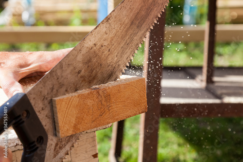 A metal hand saw with a black plastic handle with sharp teeth in the hands of a carpenter when sawing wooden beams with small chips flying to the sides. on a summer day. Construction on a cottage.