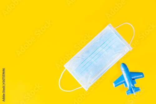 Medical face mask and blue toy plane isolated on yellow background. Travel safely on holiday flights to prevent the spread of the virus. Going vacations in summer during the coronavirus pandemic.