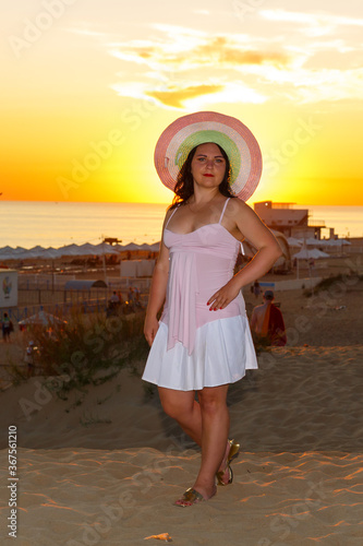 Woman in a white dress and a straw hat on the beach against the background of the sea at sunset.