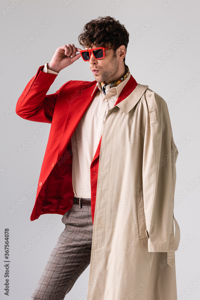 confident, stylish man touching sunglasses while looking away isolated on grey