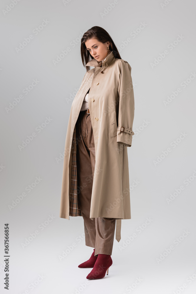 full length view of stylish girl with trench coat on shoulders posing on grey full length view of stylish girl with trench coat on shoulders posing on grey