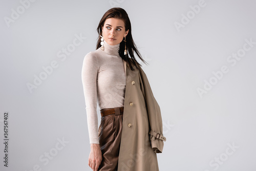 beautiful, elegant girl holding trench coat and looking away isolated on grey