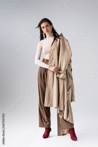 full length view of attractive, trendy girl with trench coat on shoulder looking at camera on grey
