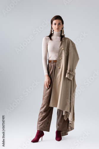 full length view of beautiful, stylish girl with trench coat on shoulder looking at camera on grey
