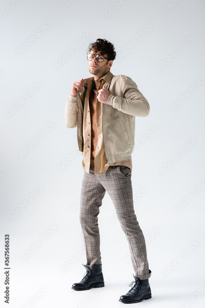 Fototapeta premium full length view of stylish man touching jacket while looking at camera on grey