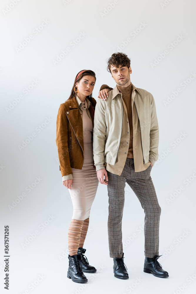 Full length view of stylish woman in autumn outfit posing near man standing with hand in pocket on grey Full length view of stylish woman in autumn outfit posing near man standing with hand in pocket on grey