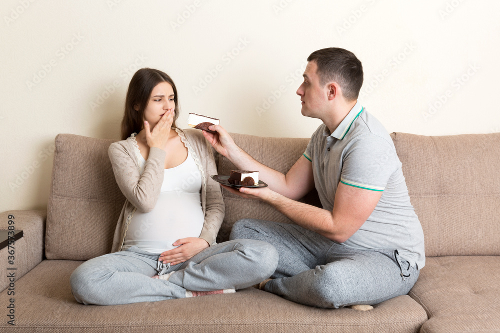 Husband offers a piece of cake to his pregnant wife but she refuses and makes stop gesture because she feels sick. Feeling bad during pregnancy concept Husband offers a piece of cake to his pregnant wife but she refuses and makes stop gesture because she feels sick. Feeling bad during pregnancy concept