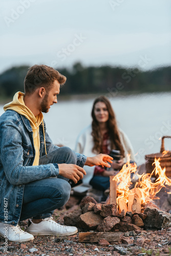 selective focus of handsome man sitting and looking at burning bonfire near girlfriend