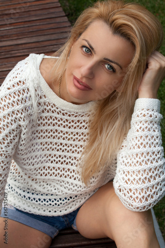 portrait of a blonde woman in a white sweater and denim shorts on a green grass background 	