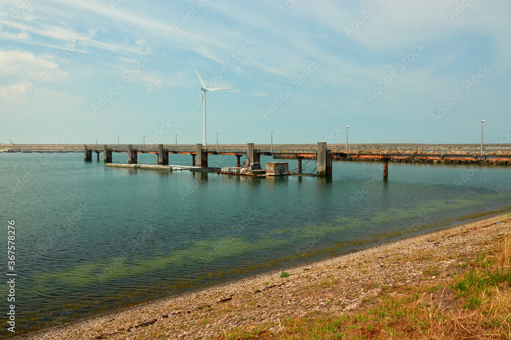 Obraz premium Brown pebble beach and deep blue water, a long pier with wooden pillars, wind turbine and dam in the background. Netherlands, Vrowenpolder.
