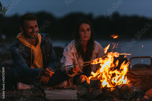 selective focus of happy couple roasting marshmallows on sticks near bonfire at night
