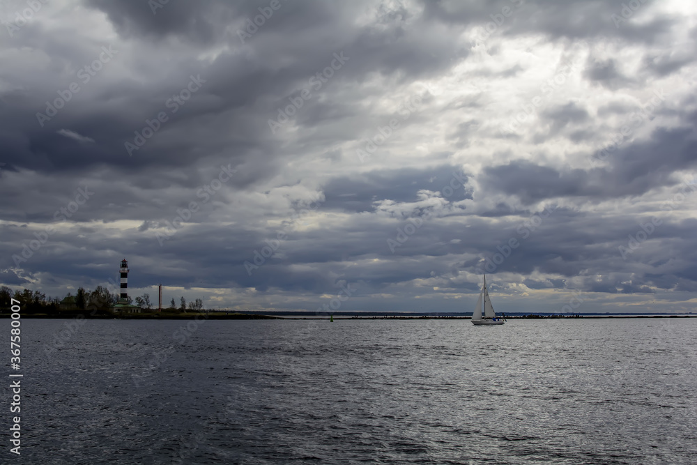 White sailboat and stripped lighthouse at place where Daugava river flows into the Baltic sea, under dramatic sky.