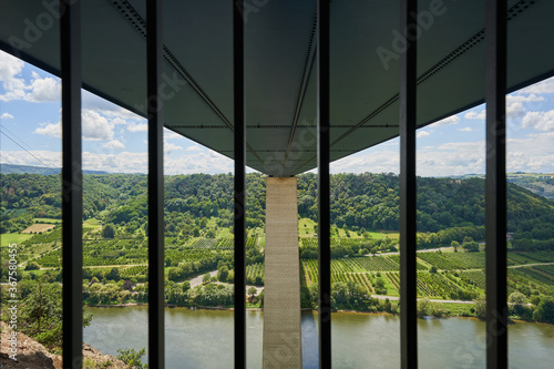 Moselle valley bridge from below, focus on gray pillar. Photographed through a grid. River and many green trees. Germany.