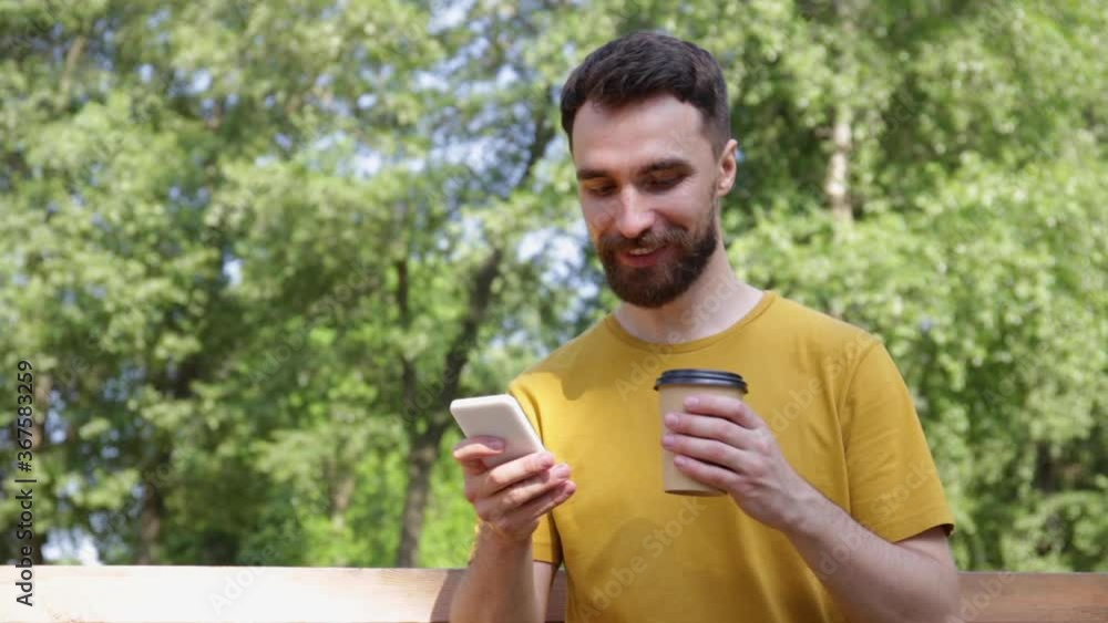 smiling man using smartphone and drinking coffee in park smiling man using smartphone and drinking coffee in park