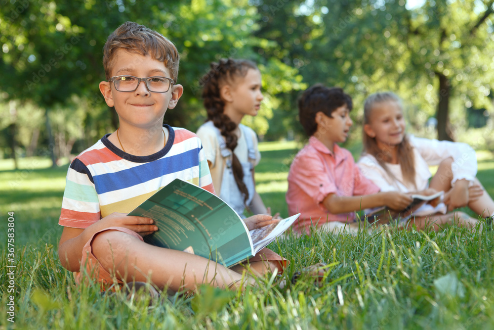 Fototapeta premium Charming little boy smiling to the camera, while reading a book, sitting on the grass