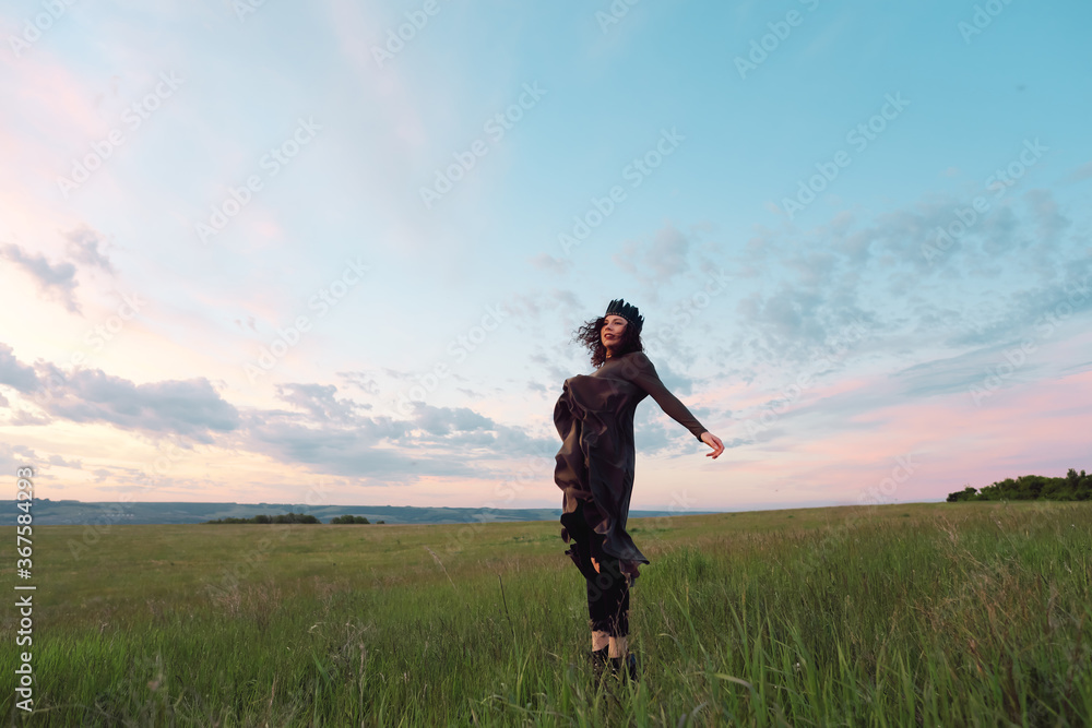 A young woman of 25-30 years old with curly red hair, in a green dress and a diadem with feathers, is spinning and dancing against the background of beautiful nature at dawn. 