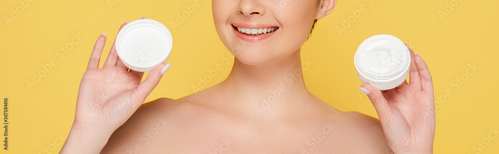 cropped view of smiling beautiful naked woman holding container with cosmetic cream isolated on yellow, panoramic shot cropped view of smiling beautiful naked woman holding container with cosmetic cream isolated on yellow, panoramic shot