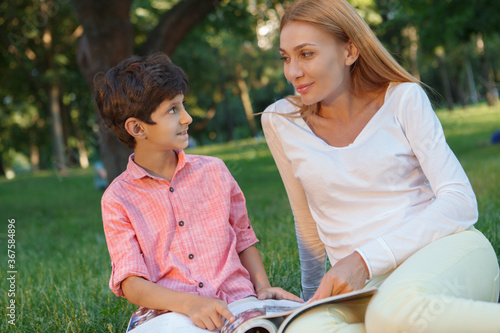 Cute happy little boy smiling at his female teacher, sitting with a book in the grass