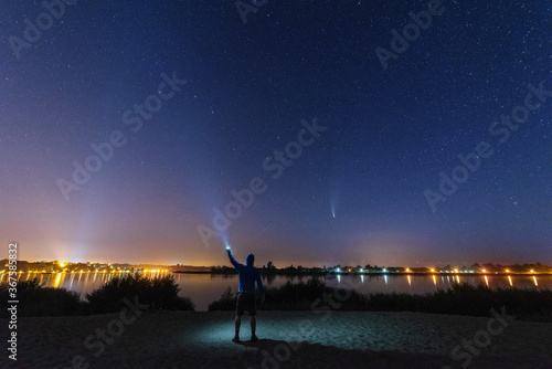 Comet NEOWISE in the night sky over the river, with the Milky Way and a tourist with a tent