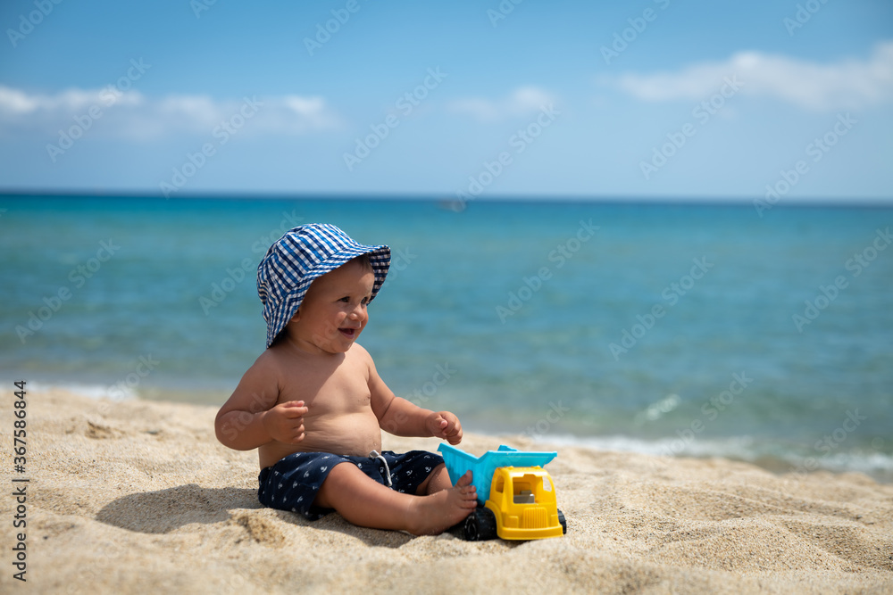 Authentic shot of happy toddler baby boy in blue vichy hat is smiling in camera while playing with a toy truck on a sandy beach during family vacation holiday at sea. Authentic shot of happy toddler baby boy in blue vichy hat is smiling in camera while playing with a toy truck on a sandy beach during family vacation holiday at sea.