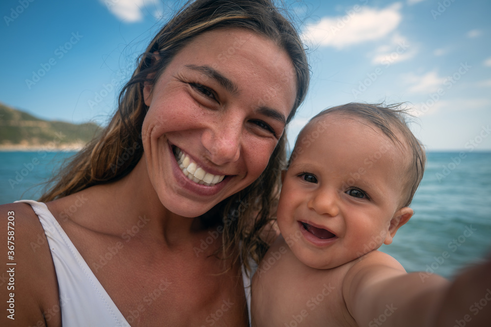 Authentic close up shot of happy carefree smiling neo mother and her newborn baby making selfie or video call to father or relatives on a beach with seaside in a sunny day. Authentic close up shot of happy carefree smiling neo mother and her newborn baby making selfie or video call to father or relatives on a beach with seaside in a sunny day.