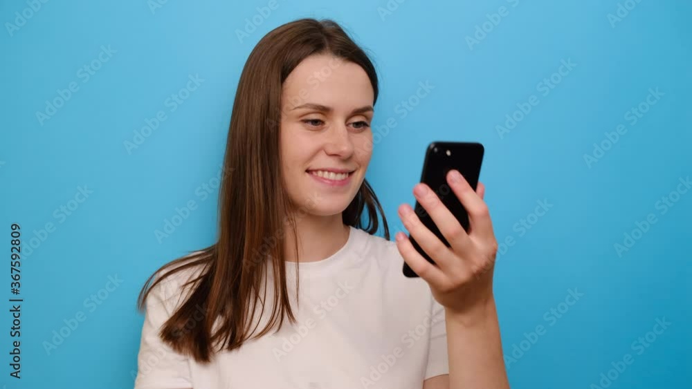 Portrait of young woman use mobile phone smile while watching video on internet, social media networks user, wears t-shirt, isolated on blue studio background. Read message feels pleased good news Portrait of young woman use mobile phone smile while watching video on internet, social media networks user, wears t-shirt, isolated on blue studio background. Read message feels pleased good news