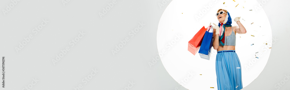 Horizontal image of positive stylish girl holding red and blue shopping bags under falling confetti near circle on white background Horizontal image of positive stylish girl holding red and blue shopping bags under falling confetti near circle on white background