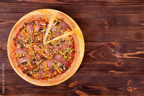 Pizza with chicken breast, corn, bacon and mushrooms, with a slice slightly removed on a wood plate which is on wooden rustic table, top view and copy space