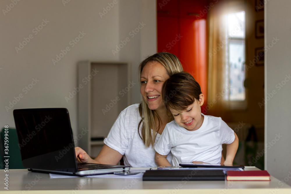 Young Caucasian single mother working on laptop while her son is using digital tablet.Mother and her son spending time together at home.Mother using laptop and her son using tablet.Technology concept. Young Caucasian single mother working on laptop while her son is using digital tablet.Mother and her son spending time together at home.Mother using laptop and her son using tablet.Technology concept.