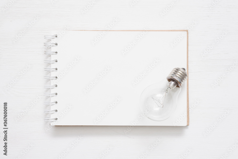 Notepad with blank pages and a lightbulb on the white wooden table background. Ideas list template. Notepad with blank pages and a lightbulb on the white wooden table background. Ideas list template.