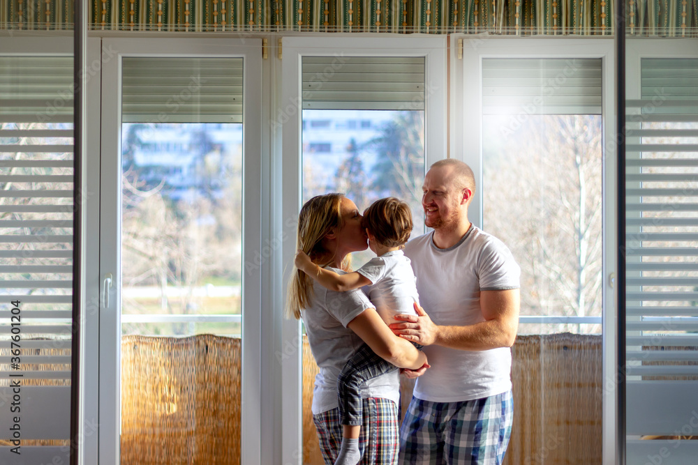 Fototapeta premium Portrait of beautiful young family standing on balcony and having fun together.Shot of happy family playing at home.Image of husband and wife in pajamas standing on balcony,and holds their son in arms