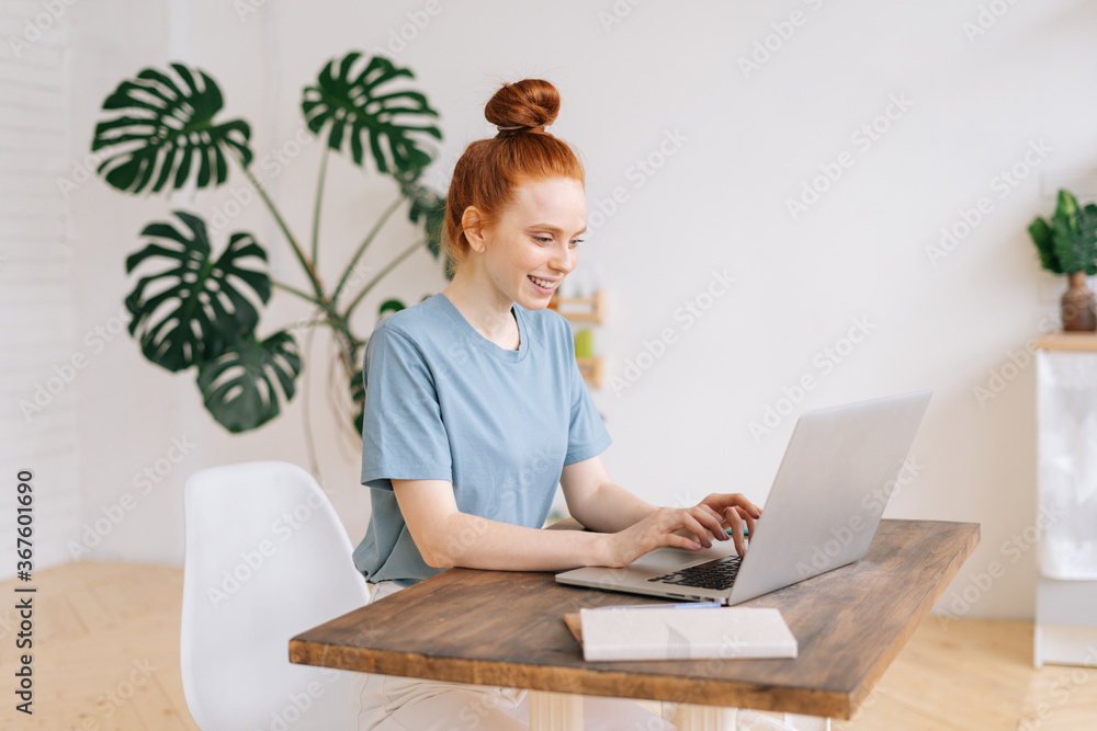 Cheerful smiling redhead young business woman is working on laptop computer at the desk in light cozy room at the home office. Cheerful smiling redhead young business woman is working on laptop computer at the desk in light cozy room at the home office.