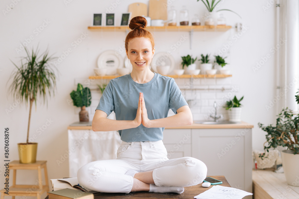 Portrait of peaceful meditating redhead young woman is putting her hands together on chest while sitting on desk at cozy home office, looking at camera. Happy female sitting in lotus pose. Portrait of peaceful meditating redhead young woman is putting her hands together on chest while sitting on desk at cozy home office, looking at camera. Happy female sitting in lotus pose.