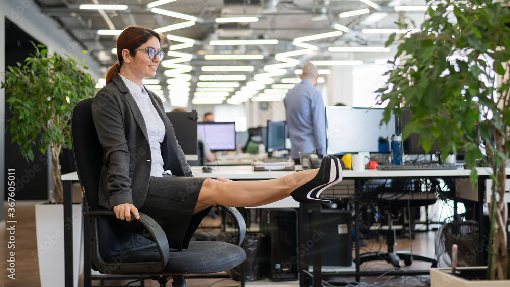 Happy business woman doing abdominal muscle exercises in an open space office. A red-haired smiling female employee in a skirt and high heels is dying off a chair in the workplace. Fitness at work. Happy business woman doing abdominal muscle exercises in an open space office. A red-haired smiling female employee in a skirt and high heels is dying off a chair in the workplace. Fitness at work.