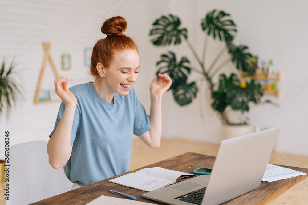 Cheerful redhead young businesswoman is working laptop and getting good message at remote work place at the desk in home office. Exited girl goes crazy because of good news from the internet. Cheerful redhead young businesswoman is working laptop and getting good message at remote work place at the desk in home office. Exited girl goes crazy because of good news from the internet.