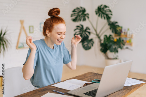 Konstfotografi Cheerful redhead young businesswoman is working laptop and getting good message at remote work place at the desk in home office