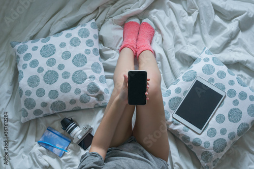 Girl in bed looking at her cell phone. Around her she has a tablet, a camera and a notebook.