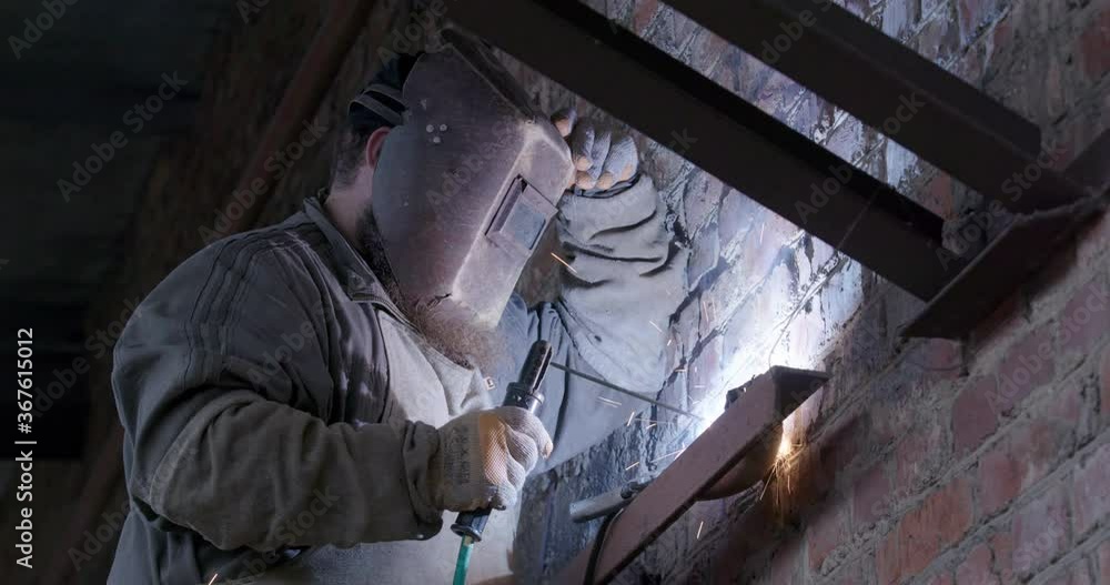 A welder makes a metal structure in an industrial building. Blacksmith welder working in a protective mask with a welding machine, bright sparks and flashes in slow motion A welder makes a metal structure in an industrial building. Blacksmith welder working in a protective mask with a welding machine, bright sparks and flashes in slow motion