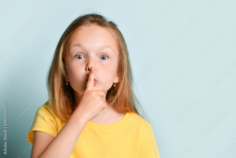 Blonde child in yellow t-shirt. She is showing be quiet sign by her forefinger while posing against turquoise studio background. Close up Blonde child in yellow t-shirt. She is showing be quiet sign by her forefinger while posing against turquoise studio background. Close up