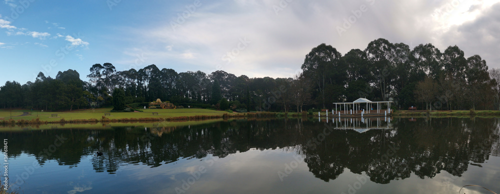 Obraz premium Beautiful morning panoramic view of a still pond in a park with stunning reflections of blue puffy sky and tall trees, Fagan park, Galston, Sydney, New South Wales, Australia