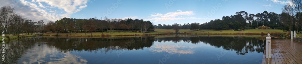 Beautiful morning panoramic view of a still pond in a park with stunning reflections of blue puffy sky and tall trees, Fagan park, Galston, Sydney, New South Wales, Australia Beautiful morning panoramic view of a still pond in a park with stunning reflections of blue puffy sky and tall trees, Fagan park, Galston, Sydney, New South Wales, Australia