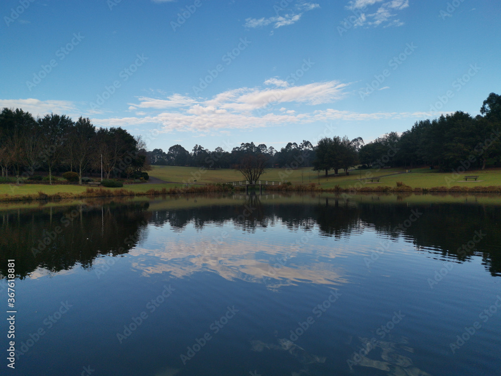 Beautiful morning view of a still pond in a park with stunning reflections of blue puffy sky and tall trees, Fagan park, Galston, Sydney, New South Wales, Australia Beautiful morning view of a still pond in a park with stunning reflections of blue puffy sky and tall trees, Fagan park, Galston, Sydney, New South Wales, Australia