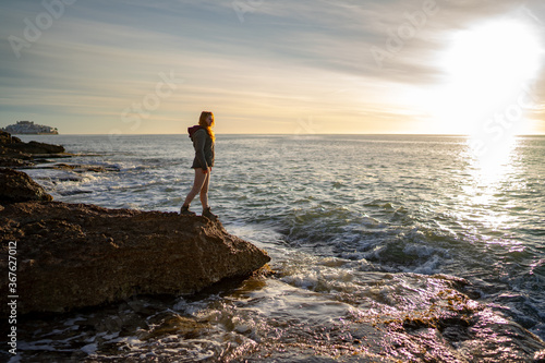 Side view of woman looking at seascape against sky while standing on rock during sunset